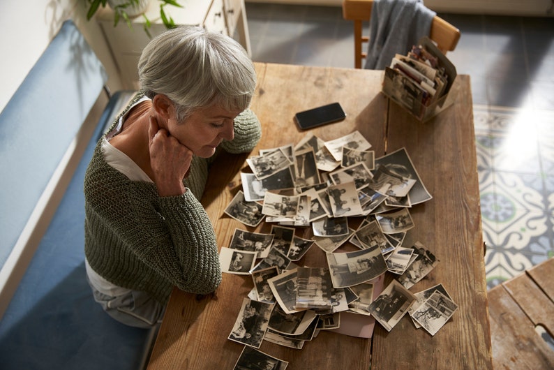 May include: A woman sits at a wooden table, looking through a pile of black and white photographs. The photos are scattered across the table, and the woman is holding one in her hand.
