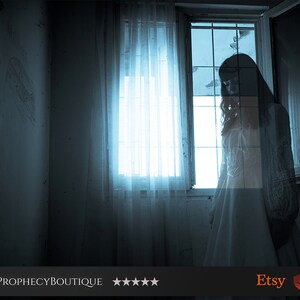 May include: A woman in a white dress stands by a window in a dimly lit room. The window is covered with a sheer curtain and the light from outside casts shadows on the wall.
