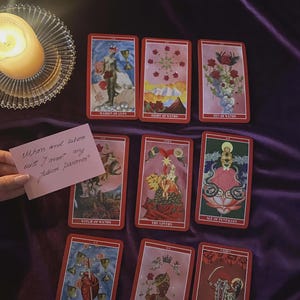 May include: A collection of tarot cards with red borders and various illustrations, including the Maiden of Cups, Eight of Wands, and Death. A lit candle sits in a glass holder. A handwritten note asks, "When and where will I meet my future partner?"
