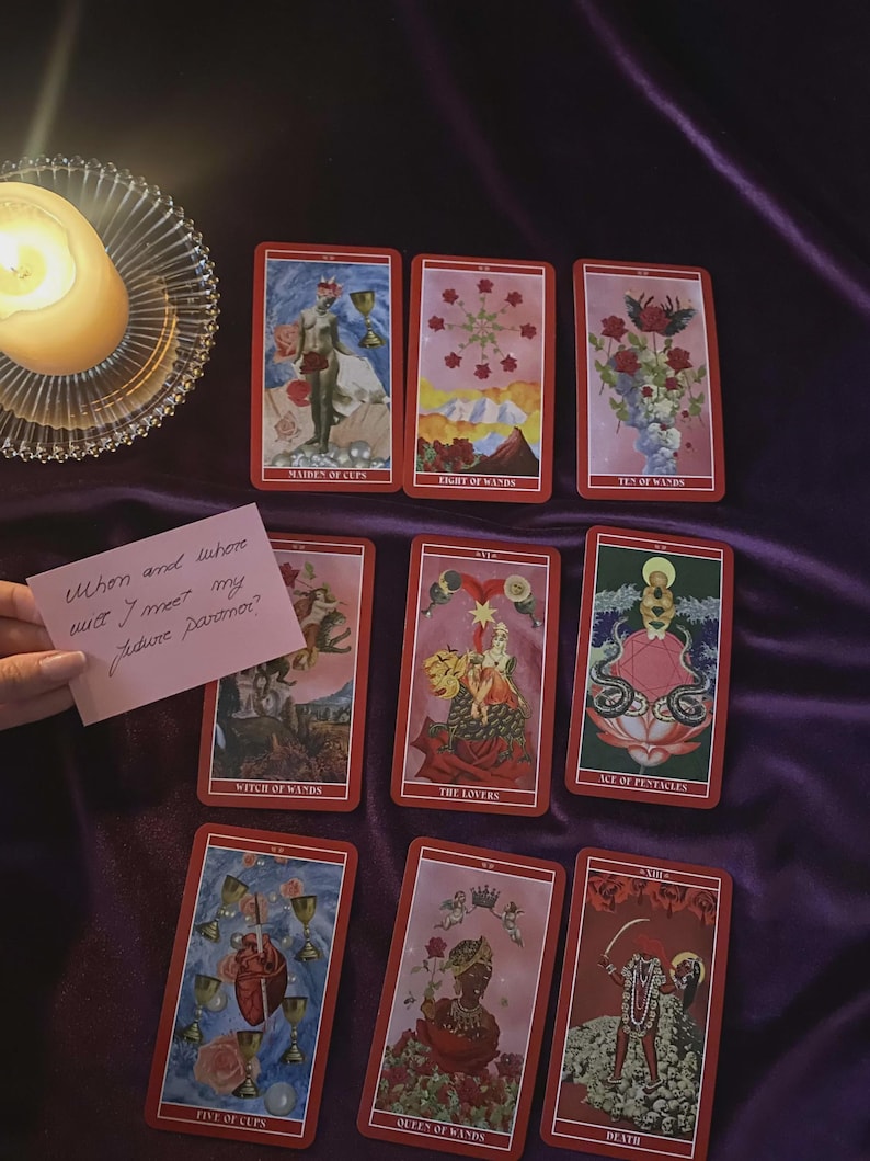 May include: A collection of tarot cards with red borders and various illustrations, including the Maiden of Cups, Eight of Wands, and Death. A lit candle sits in a glass holder. A handwritten note asks, "When and where will I meet my future partner?"