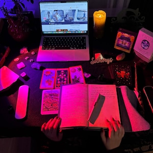 May include: A laptop, tarot cards, crystals, and a lit candle are arranged on a dark surface. The laptop displays a website. A journal is open with a pen resting on the pages. The scene is illuminated with pink and purple lighting, creating a mystical ambiance.