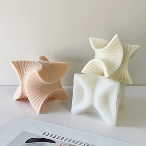 May include: Three geometric candles in a white, pink, and cream color scheme. The candles are stacked on top of each other and have a unique, folded design.