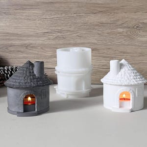 May include: Three ceramic house-shaped candle holders in grey and white, with lit candles inside. A white cylindrical object is in the centre. The candle holders have detailed roof and chimney designs, creating a cosy, decorative ambience.