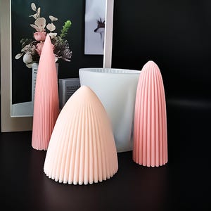 Pode incluir: Uma coleção de moldes de velas cor-de-rosa em várias formas e tamanhos. Os moldes têm um design canelado e estão dispostos numa superfície preta. Também está presente um molde cilíndrico branco, juntamente com uma imagem emoldurada e um vaso de flores.