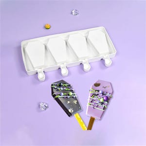 Może przedstawiać: Biała forma do lodów w kształcie trumny z czterema przegródkami, obok dwóch zdobionych lodów na patyku. Jedna lodowa jest ciemna z zieloną i fioletową posypką, druga jest jasnofioletowa z podobnymi dekoracjami. Lody na patyku są na żółtych i brązowych patyczkach.