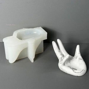 Può includere: Uno stampo in silicone bianco a forma di mano con una piccola ciotola nel palmo. Accanto allo stampo c'è un piatto bianco a forma di mano con un design marmoreo grigio.