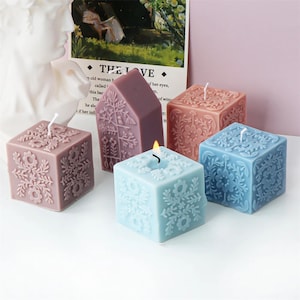 May include: Four decorative square candles with floral designs. The candles are in shades of blue, pink, and purple. One candle is shaped like a house.