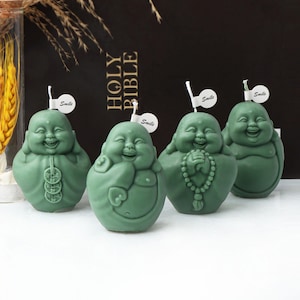 May include: Four green smiling Buddha candles with the word "Smile" on each. The candles are sitting on a black surface with a book that says "Holy Bible" in the background.