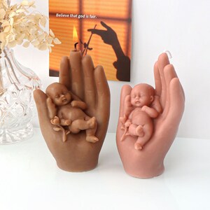 May include: Two hand-shaped candles, each holding a sleeping baby. One candle is brown and the other is pink.