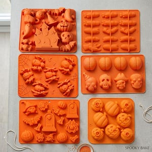 Puede incluir: Seis moldes de silicona naranja para hornear con diversas formas temáticas de Halloween, incluyendo fantasmas, calabazas, calaveras, arañas, murciélagos, lápidas y dedos. El texto SPOOKY BAKE está impreso en la esquina inferior derecha.