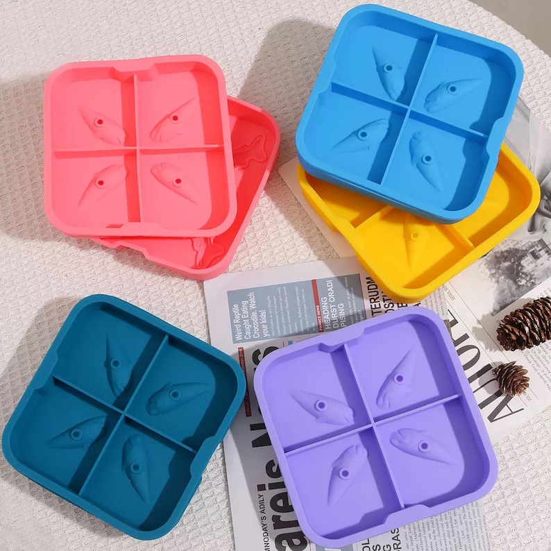 May include: A collection of colorful silicone ice cube trays in pink, blue, yellow, teal, and purple. Each tray is divided into four sections, with a unique design in each. The trays are arranged on a patterned surface with a newspaper and pinecone.