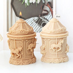 Puede incluir: Dos velas de color beige en forma de carrusel con detalles intrincados y un diseño de caballo. La vela de la izquierda tiene una mecha encendida. La vela de la derecha tiene una pequeña etiqueta blanca con la palabra "Smile" escrita en ella.