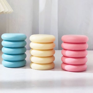 Op de afbeelding: Drie stapels ronde, decoratieve kaarsen in verschillende kleuren. De stapels zijn lichtblauw, crème en roze. Elke stapel heeft vijf kaarsen.