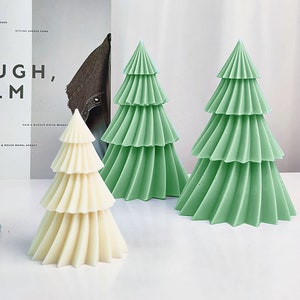 Puede incluir: Tres velas decorativas con forma de árbol de Navidad. Una es de color blanco roto y las otras dos son de color verde claro. Cada vela tiene un diseño en capas, que se asemeja a las ramas de un árbol. Las velas están colocadas sobre una superficie blanca.