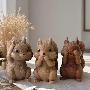 May include: Three brown squirrel figurines in various poses: covering ears, mouth, and eyes. The figurines are in shades of brown and tan, with detailed features. The squirrels are on a white surface, with dried plants in the background.