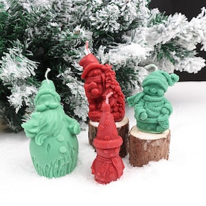 May include: Four decorative wax candles shaped like holiday figures including a light green Santa Claus, a red figure with a tree, a small red snowman with a snowflake hat, and a green snowman wearing a knit sweater and scarf, each with a wick.
