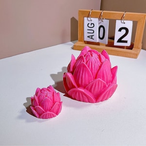 May include: Two pink lotus flower shaped candles, one larger than the other, on a white surface. The larger candle is in the foreground and the smaller candle is in the background. The date August 02 is visible on a wooden calendar in the background.