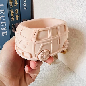 May include: A light pink ceramic planter shaped like a vintage Volkswagen van. The planter has a rounded top and a rectangular base. The van has a detailed design with windows, wheels, and a front grill.