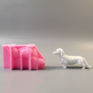 Puede incluir: Un molde de silicona rosa con forma de perro salchicha, junto a una figura de perro salchicha blanco. El molde tiene forma rectangular con soportes internos. La figura es detallada, mostrando el pelaje y las características del perro.