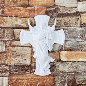May include: A white religious wall cross featuring a sculpted figure with outstretched arms. The cross has the letters INRI at the top. The cross is mounted on a brick-patterned wall.