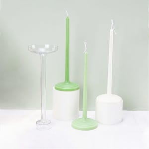 May include: Three tall candles on white pedestals. Two candles are green and one is white. The candles are on a white surface.
