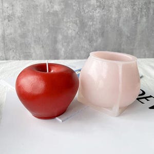 May include: Two candle molds, one shaped like an apple and the other a round pot. The apple mold is red and the pot mold is pink.