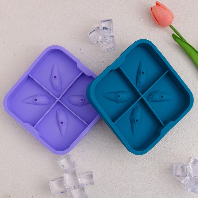 May include: Two silicone ice cube trays, one purple and one teal, each with four molds shaped like stylized teeth. Clear ice cubes are scattered around the trays, and a pink tulip is in the background. The trays are square with rounded corners.