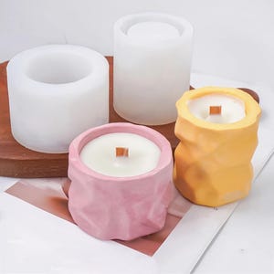 Puede incluir: Tres moldes para velas, dos son de silicona blanca y uno es rosa con un acabado texturizado. Los moldes de silicona son cilíndricos y el molde rosa tiene una forma más orgánica. Hay dos velas en los moldes, una es amarilla y otra es rosa.