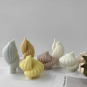 Op de afbeelding: Een verzameling decoratieve, abstracte sculpturen in de vorm van knoflookbollen. De sculpturen zijn in verschillende pastelkleuren, waaronder geel, lavendel en wit. De vormen hebben een geribbelde textuur en zijn op een wit oppervlak geplaatst.