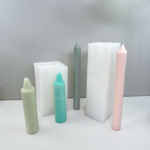 May include: An assortment of candles in various colors and shapes. The candles include a light green, teal, gray, and pink candle. There are also two white, rectangular candle molds. The candles are on a white surface with a gray background.