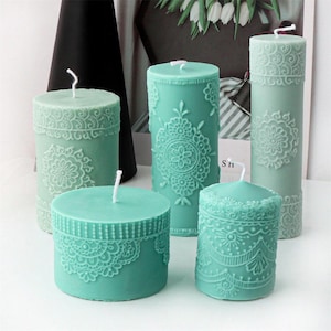 Può includere: Una collezione di candele a colonna color turchese con intricati motivi floreali e in pizzo. Le candele variano in altezza e forma, con stoppini bianchi. I disegni sono in rilievo sulle superfici delle candele, creando un effetto strutturato. Le candele sono disposte su una superficie bianca.