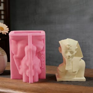 May include: A pink silicone mold with the word "WOOOO" and a bust of a figure, next to a cream-colored bust of an Egyptian pharaoh. The pharaoh bust is on a square base. Both items are on a wooden surface.