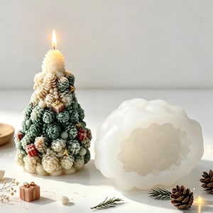 Puede incluir: Una vela encendida con forma de árbol de Navidad hecha de piñas apiladas en tonos crema y verde salvia, decorada con pequeños regalos rojos y estrellas doradas, junto a un molde de silicona blanco translúcido con la misma forma de árbol.
