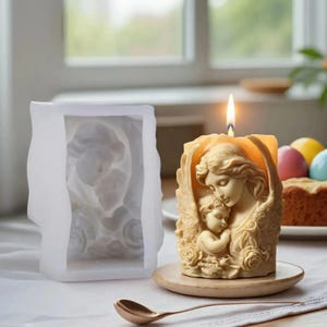 May include: A lit beige candle with a detailed relief of a mother and child, set on a wooden coaster. A white silicone mold of the same design sits to the left. A lit flame sits atop the candle.