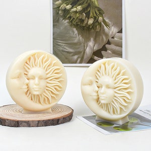 May include: Two white ceramic sculptures of a sun and moon with faces. The sculptures are on wooden bases and are displayed on a white surface.
