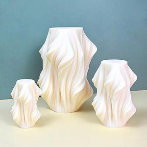 Puede incluir: Tres candelabros abstractos de color blanquecino con un diseño de tela drapeada. Los candelabros varían en tamaño, siendo el más grande el del centro. El diseño similar a la tela tiene una textura ondulada, creando una apariencia suave y fluida. El fondo es azul claro.