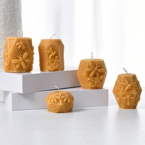 May include: Five hexagonal beeswax candles in different sizes, each featuring a honeycomb and bee design. The candles are a warm yellow colour and are displayed on a white surface. The designs include bees, flowers, and honeycomb patterns.