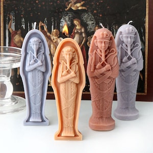May include: Four decorative candles shaped like Egyptian sarcophagi. The candles are in shades of purple, pink, and brown.