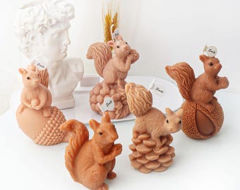 May include: Five brown squirrel-shaped candles with different acorn-shaped bases. The squirrels are sitting upright with their tails curled behind them. The bases are decorated with different textures, including a pine cone, a smooth acorn, and a textured acorn.