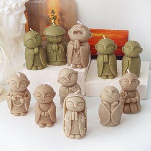 May include: A set of ten small, hand-crafted, brown and green candle sculptures of Buddha figures in various poses. The candles are arranged on a white surface.