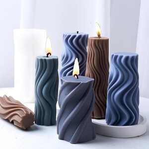 K&ouml;nnte beinhalten: F&uuml;nf dekorative Kerzen mit einem wellenf&ouml;rmigen Design. Die Kerzen sind in den Farben Braun, Blau und Grau gehalten. Die Kerzen sind angez&uuml;ndet und die Flammen sind sichtbar.