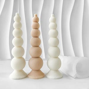 Può includere: Tre candele a colonna decorative in avorio e beige. Le candele hanno un design impilato e sferico con dettagli a coste verticali. Le candele a sinistra e a destra sono avorio, mentre la candela centrale è beige. Uno stampo bianco e flessibile è sullo sfondo.