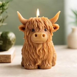 May include: A tan-colored candle shaped like a Highland cow with a lit wick. The candle has detailed fur, horns, and a cute face. The candle is a decorative item.