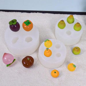 May include: A collection of miniature fruit sculptures, including pears, oranges, and a peach, alongside silicone molds. The fruits are brightly colored and detailed, with green stems and leaves. The molds are white and circular, with multiple cavities.