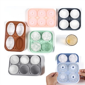 May include: Assortment of ice cube molds in various shapes, including footballs, soccer balls, baseballs, golf balls, and round shapes. The molds are in shades of brown, pink, green, gray, and blue. A glass of liquid is also present.