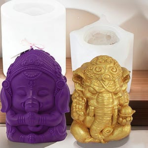 May include: A purple and gold Ganesh statue. The purple statue is in a seated position with hands together in prayer. The gold statue is also in a seated position. Two white silicone molds are in the background.