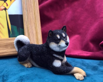 May include: Black and white Shiba Inu dog figurine lying down on a blue surface. The figurine is detailed with black fur and white markings.