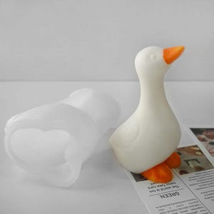 Könnte beinhalten: Eine weiße Gänsefigur mit orangefarbenem Schnabel und Füßen. Daneben befindet sich eine weiße Silikonform in Herzform. Die Gans steht auf bedrucktem Papier.