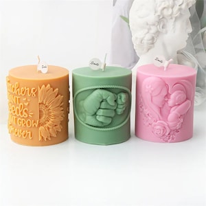 Può includere: Tre candele a pilastro con diversi design. La prima candela è arancione e ha il testo "Teachers plant seeds that grow forever". La seconda candela è verde e ha un design con un'impronta di mano. La terza candela è rosa e ha un design con una madre e un bambino.