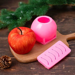May include: A realistic red apple sits beside a pink sphere-shaped mould and a pink rectangular mould with curved lines. The items are on a wooden chopping board, with a pinecone and evergreen branches in the background.
